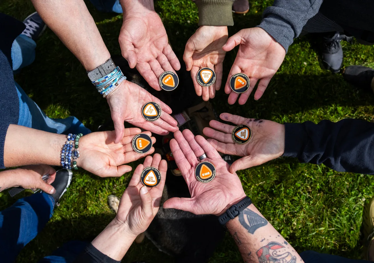 A circle of veterans’ open hands, each holding the same guitar-pick coin